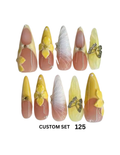 A set of 10 custom false press-on nails. The nails feature a peach-to-yellow gradient, yellow floral 3D designs, white feather designs, and glittery gold butterfly accents.