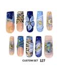 A set of 10 custom press-on nails.  The nails feature various designs including blue and peach gradients, floral details, gold chain accents, and celestial motifs like moons and stars.